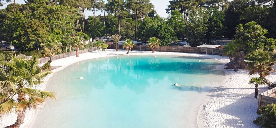 A panoramic lagoon with a white beach lined with lounge chairs, palm trees and forest under a blue sky