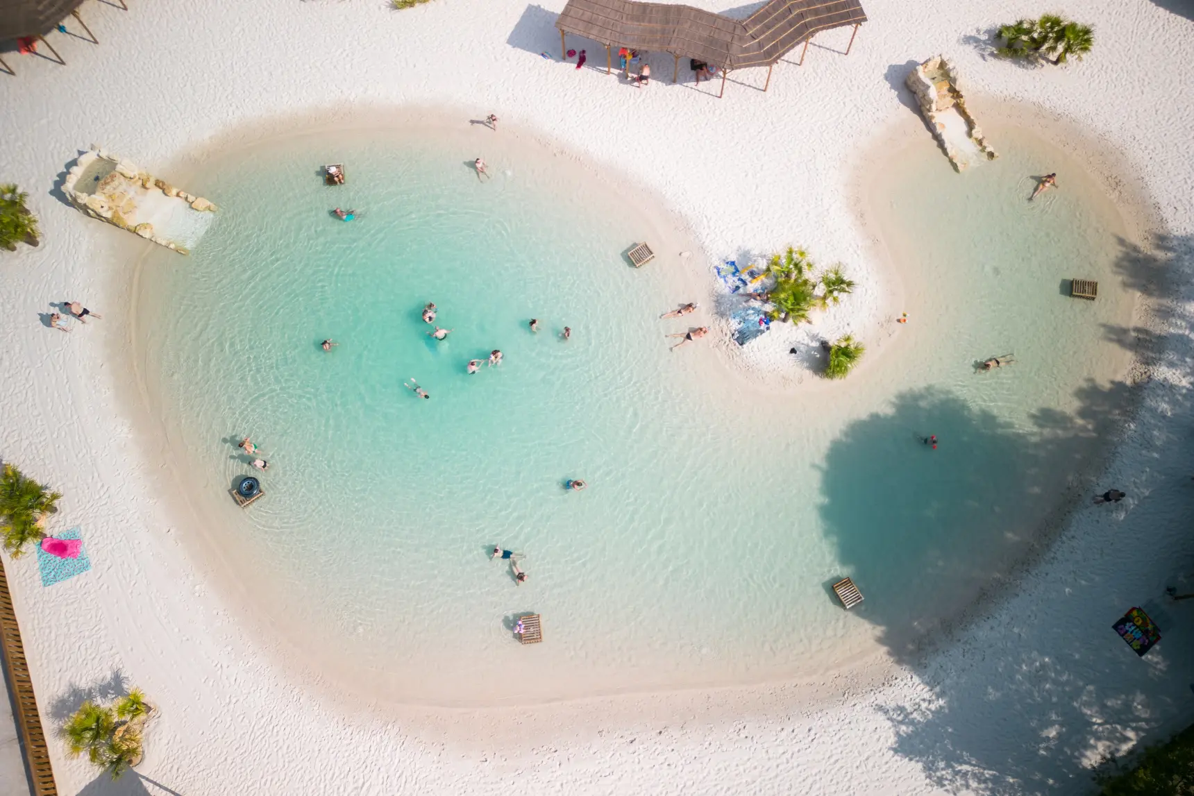 Aerial view of a multi-zone lagoon with white sand beaches at a campground, organized seating amid green forest