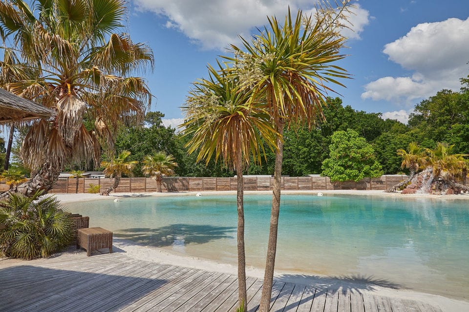 A luxury lagoon with turquoise water and white sand, a wood deck with lounge seating, palm trees and forest behind