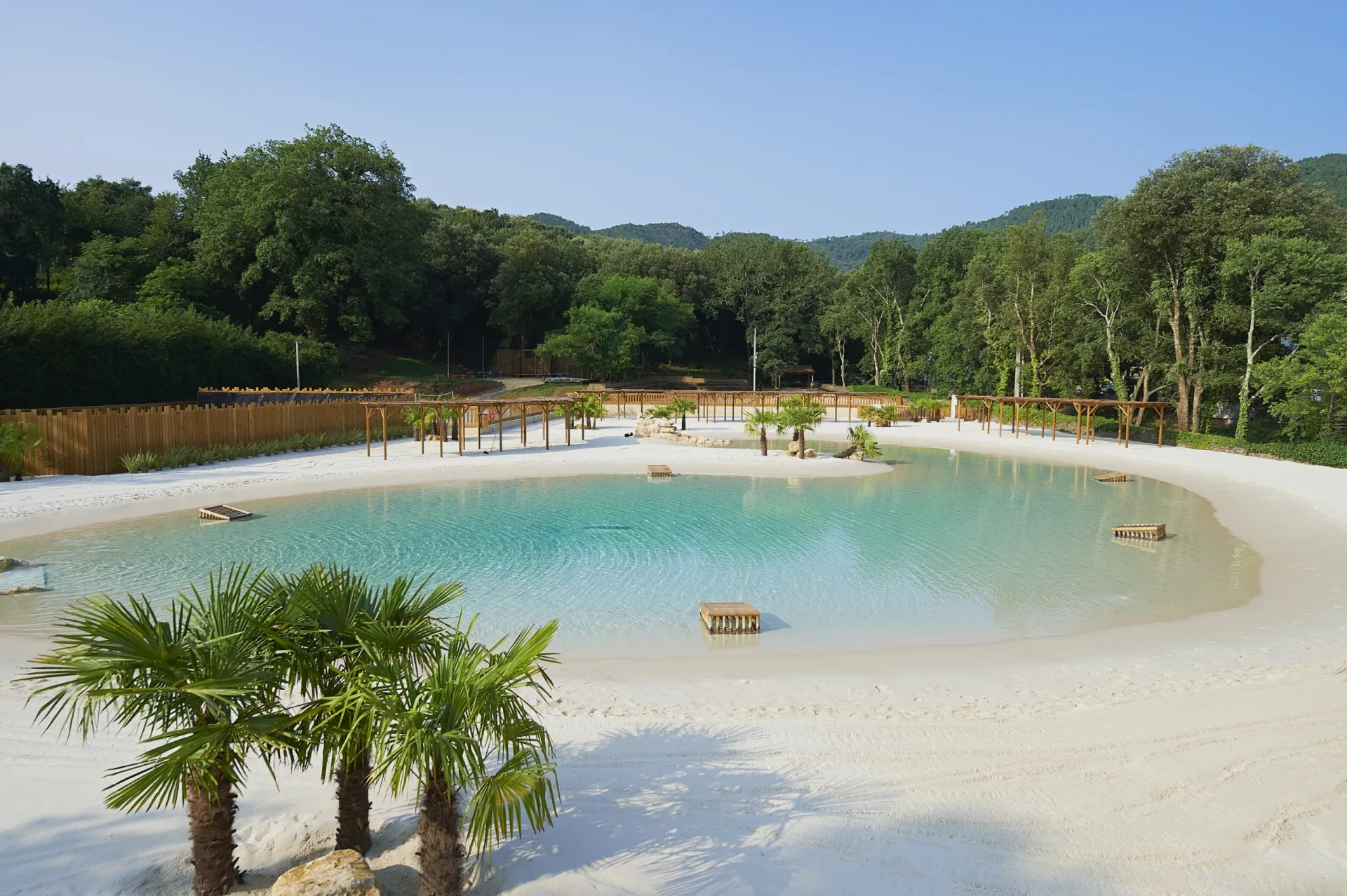 A finished lagoon reading as the outcome of a build: turquoise water, warm fine sand, natural rock, integrated planting, sun-drenched and composed